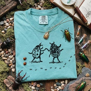 May include: A light teal Comfort Colors t-shirt featuring a black graphic of two dancing beetles and a line of ants. A gold necklace with a scarab beetle pendant and several decorative beetles and vials are also present.