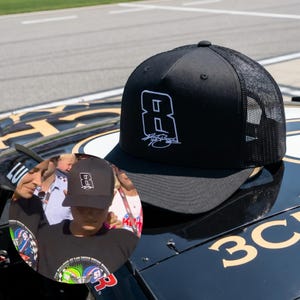 May include: Black trucker hat featuring a white number 8 and a signature. The hat has a mesh back and a curved brim. The hat is sitting on a black surface with gold lettering.
