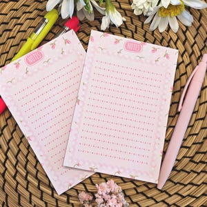 Op de afbeelding: Twee roze notitieblokken met bloemen- en parelrandontwerpen, elk met het woord 'NOTES' bovenaan. De notitieblokken liggen op een geweven oppervlak, met een roze pen en een gele pen in de buurt. Witte bloemen en roze bloemen zijn ook aanwezig.