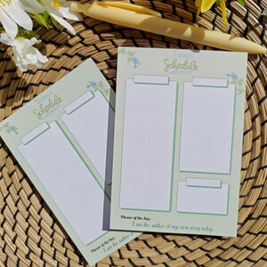 May include: Two mint green schedule notepads with the text "Today's Schedule" and a phrase of the day. Each notepad has a clipboard design with blank spaces for notes. A yellow pen and flowers are in the background.