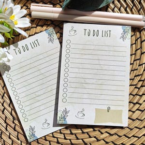 Könnte beinhalten: Zwei beige "TO DO LIST" Notizblöcke mit linierten Seiten, Kaffeetassen-Illustrationen und botanischen Akzenten. Zwei Stifte liegen über den Notizblöcken. Die Notizblöcke liegen auf einer gewebten Oberfläche.