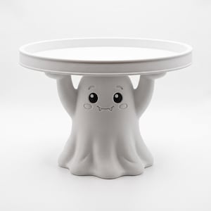 Pu&ograve; includere: Un'alzata per torte bianca con una base a forma di fantasma scolpito, le cui braccia sollevate sostengono un vassoio circolare. Il fantasma ha un volto amichevole con grandi occhi neri, sopracciglia, guance rosate e un sorriso.