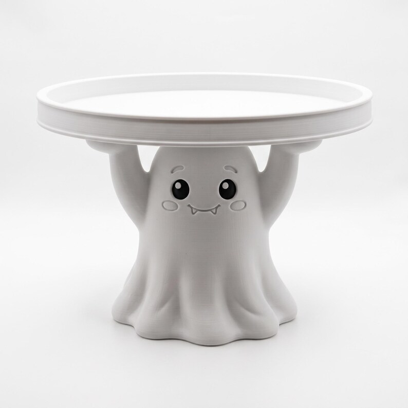 Pu&ograve; includere: Un'alzata per torte bianca con una base a forma di fantasma scolpito, le cui braccia sollevate sostengono un vassoio circolare. Il fantasma ha un volto amichevole con grandi occhi neri, sopracciglia, guance rosate e un sorriso.