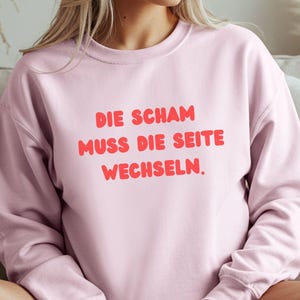 Könnte beinhalten: Hellrosa Sweatshirt mit dem deutschen Text "DIE SCHAM MUSS DIE SEITE WECHSELN" in roten, fetten Buchstaben. Das Sweatshirt hat einen Rundhalsausschnitt und scheint aus einem weichen Material zu sein.
