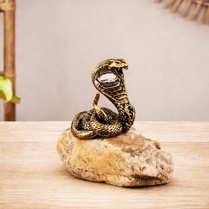 Puede incluir: Una estatua de cobra de bronce, enroscada sobre una piedra natural. La serpiente se representa con su capucha levantada, mostrando detalles intrincados. La estatua tiene un acabado metálico y es un objeto decorativo.