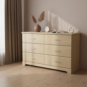 May include: A light wood six-drawer dresser with silver-toned handles. The dresser is in a room with a light brown wall, a window with a brown curtain, and a light wood floor. A vase with dried flowers, a clock, and a book are on top.