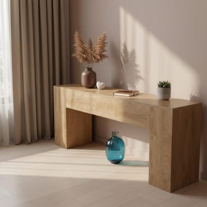 May include: A light wood console table with a rectangular design. The table is decorated with a blue glass vase, a small potted plant, a brown vase with dried flowers, a small white heart, and a notebook. The room has beige curtains and light-colored wood flooring.