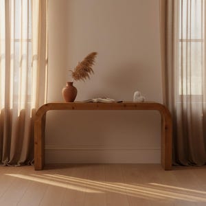 May include: A wooden console table with a curved design. A terracotta vase with dried pampas grass sits on the table, along with an open book and a small white heart-shaped sculpture. The room has beige curtains and light-colored wood flooring.