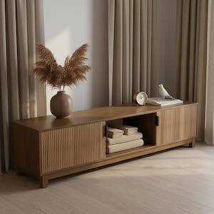 May include: A light brown wooden media console with a slatted design on the doors. The console has an open shelf with folded towels. A vase with dried plants, a clock, and a decorative bird are on top.