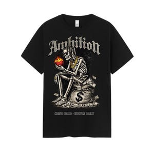 May include: Black t-shirt featuring a skeleton holding a flaming heart, seated on a money bag with a dollar sign. The word "Ambition" is above the skeleton, and the text "Grind Hard - Hustle Daily" is below.