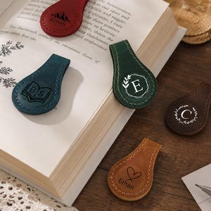 May include: Five teardrop-shaped leather magnetic bookmarks in red, blue, green, dark brown, and tan. Each features unique engravings: a mountain with the name Ethan, an open book with initials BS, a letter E in a wreath, a letter C in a wreath, and a heart with the name Ethan.