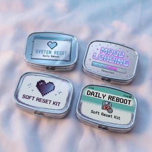Op de afbeelding: Vier rechthoekige zilveren metalen blikjes met afgeronde hoeken. Elk deksel heeft pixel art ontwerpen: één met SYSTEM RESET Daily Reboot en een hartje, één met MOOD LOADING Please Wait, één met SOFT RESET KIT en een hartje, en één met DAILY REBOOT Soft Reset Kit met een diskette-icoon.