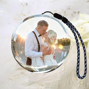 Peut inclure: Une décoration en verre transparent et ronde présentant une photographie imprimée d'un couple qui s'embrasse. La personne à gauche porte une chemise blanche avec des bretelles marron, et celle à droite une robe de mariée en dentelle blanche. Une dragonne tressée noire et bleue est fixée au sommet.