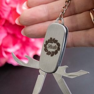 Peut inclure: Un porte-clés multifonction argenté avec plusieurs outils pliables. Le porte-clés porte le nom "Jānis" gravé dans une couronne décorative. Les outils comprennent un couteau, un ouvre-bouteille et une lime à ongles. Le porte-clés est tenu par une personne.