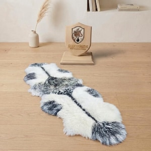 May include: A sheepskin rug with a unique shape, featuring a mix of white, gray, and black fur. The rug is placed on a wooden floor. A wooden shield with a logo and text is in the background.