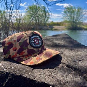 May include: A camouflage pattern baseball cap with a patch that reads "SHEEPDOG CONSERVATION". The cap is resting on a dark gray rock with a lake and trees in the background. The cap has a brown, red, and green pattern.