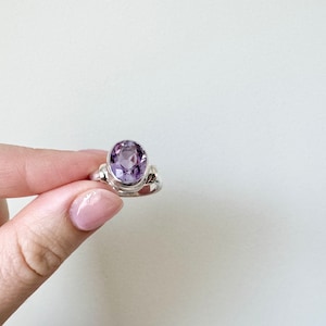 May include: A silver ring featuring a large, oval-cut amethyst gemstone. The ring's band has decorative details on the sides, and the amethyst displays a vibrant purple hue. The ring is held up against a plain white background.
