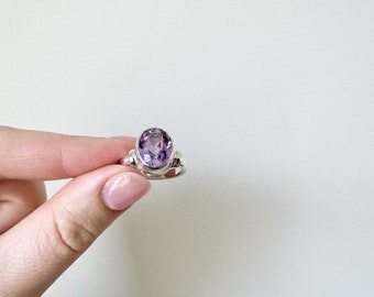 May include: A silver ring with a large, oval amethyst gemstone. The ring's band has decorative side details, and the amethyst has a vibrant purple colour. The ring is held against a plain white background.