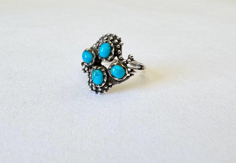 May include: A silver ring featuring five turquoise-colored stones. The ring has a detailed, textured design around the stones and band. The turquoise stones are oval-shaped and set in a cluster pattern.