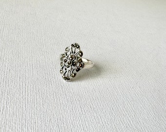 May include: A silver ring with a delicate, intricate design featuring a cluster of dark gray marcasite stones.
