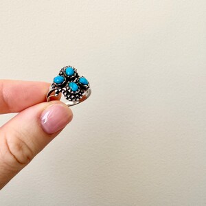 May include: A silver ring featuring five turquoise-colored stones. The ring has a detailed, ornate design with the stones clustered together. The ring is held up by a person's fingers against a neutral background.