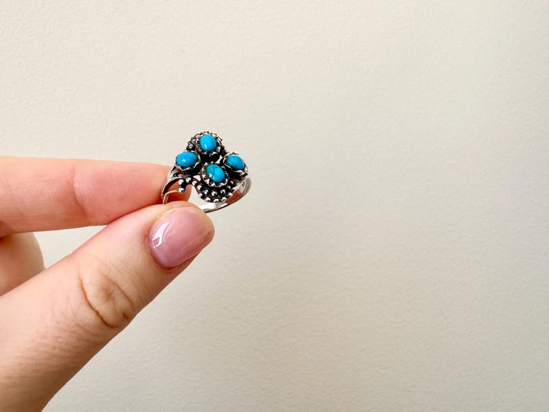 May include: A silver ring featuring five turquoise-colored stones. The ring has a detailed, ornate design with the stones clustered together. The ring is held up by a person's fingers against a neutral background.