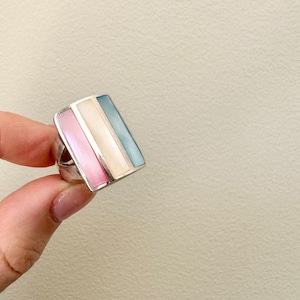 May include: A silver ring featuring a rectangular stone. The stone is composed of three layers of different coloured mother of pearl: pink, white, and blue.
