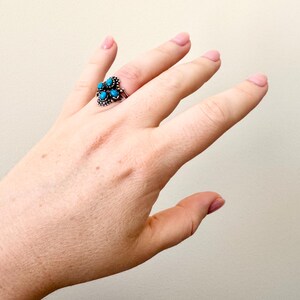 May include: A silver ring with a cluster of turquoise-colored stones. The ring is set on a hand with a light skin tone, the ring is the focal point of the image. The ring's design features multiple small, round turquoise stones.