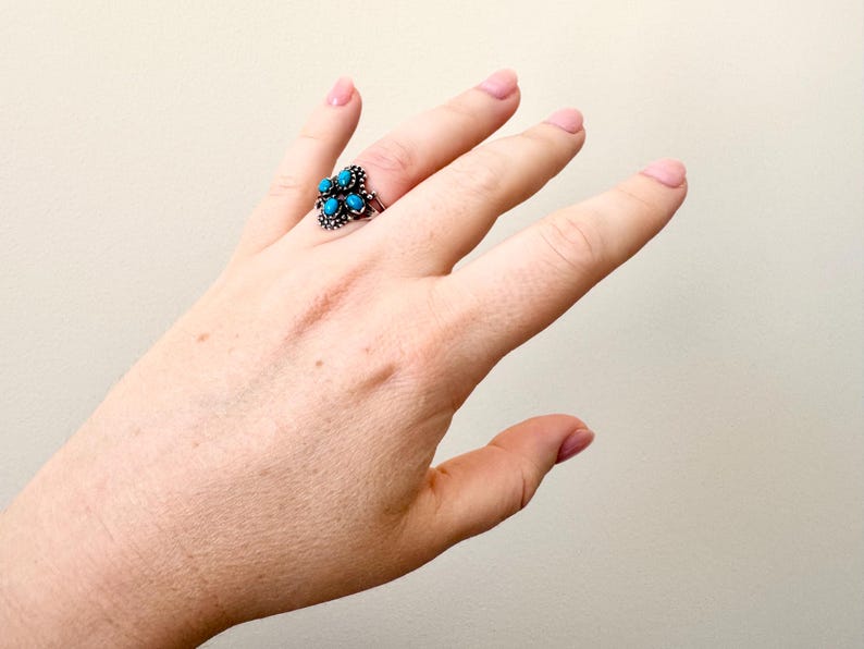 May include: A silver ring with a cluster of turquoise-colored stones. The ring is set on a hand with a light skin tone, the ring is the focal point of the image. The ring's design features multiple small, round turquoise stones.