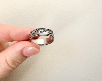 May include: A silver ring with a wave design. The ring is wide and has a textured surface.