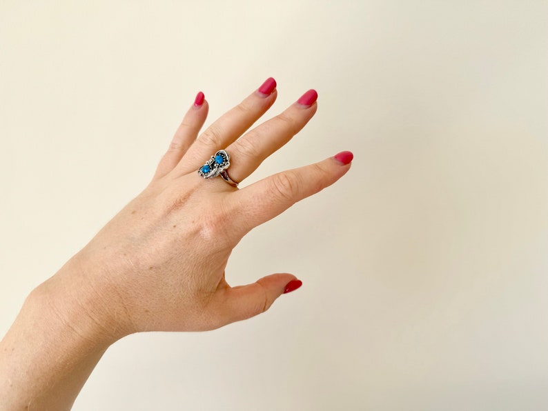 May include: A silver ring with three blue turquoise stones set in a decorative design. The ring is worn on the middle finger of a hand with pink nail polish.