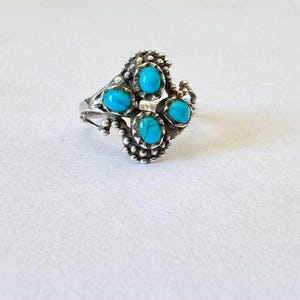 May include: A silver ring featuring four oval turquoise stones. The ring has a detailed, textured design with small beaded accents around the stones and along the band. The ring is set against a white background.