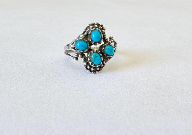 May include: A silver ring featuring four oval turquoise stones. The ring has a detailed, textured design with small beaded accents around the stones and along the band. The ring is set against a white background.
