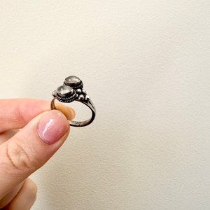 May include: A silver ring with two white stones set in the centre. The ring has a beaded design around the stones.