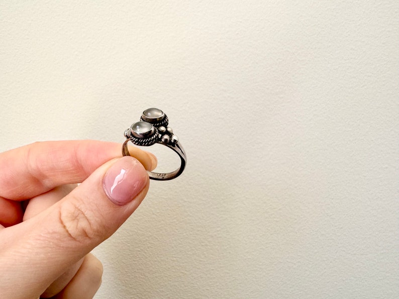 May include: A silver ring with two white stones set in the centre. The ring has a beaded design around the stones.