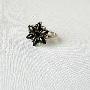 May include: A silver ring featuring a flower design with black gemstone petals. The ring has a band and small silver beads around the center of the flower. The ring is set against a white textured background.