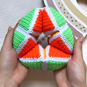 Könnte beinhalten: Ein mehrfarbiges Fidget-Spielzeug, das in Händen gehalten wird. Das Spielzeug hat abwechselnde Abschnitte in Grün, Weiß und Orange, wodurch ein ansprechendes Design entsteht. Die Struktur des Spielzeugs ermöglicht eine flexible Bewegung.