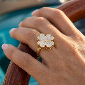 Op de afbeelding: Een gouden ring met een klaverbladontwerp met vier bladeren. De klaver is bezet met kleine, sprankelende steentjes. De ring zit aan een hand, met een houten oppervlak en een blauwe achtergrond.