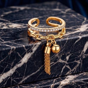 Könnte beinhalten: Ein goldfarbener offener Ring mit einem Band, das mit kleinen klaren Steinen besetzt ist, und einem zweiten Band in Form einer Gliederkette. Eine kleine goldene Kugel und eine kurze, feine goldene Kettenquaste hängen an der Kettenglied-Sektion.