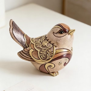 Puede incluir: Figurita decorativa de pájaro con detalles intrincados. El pájaro de cerámica presenta un cuerpo beige con detalles en marrón, dorado y burdeos. Las alas y el cuerpo están adornados con patrones ornamentados en relieve. El pájaro está posado sobre una superficie blanca.