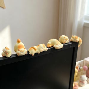 Puede incluir: Una colección de pequeñas figuras con forma de gato en varias poses, posadas sobre un monitor de ordenador negro. Las figuras son principalmente de color crema con marcas naranjas, creando una exhibición lúdica y decorativa.