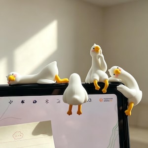 Puede incluir: Un conjunto de figuras blancas con forma de ganso, con patas y picos amarillos. Las figuras están en varias poses, algunas tumbadas, otras sentadas o inclinadas. Se colocan encima de un dispositivo electrónico negro.