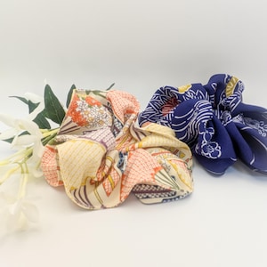 Könnte beinhalten: Zwei Stoff-Haargummis im Bild. Eines ist eine Mischung aus Creme, Orange und Gelb mit einem Blumenmuster. Das andere ist marineblau mit einem weißen Wellenmuster. Beide befinden sich auf weißem Hintergrund mit weißen Blumen.