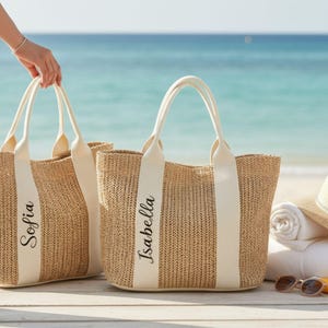 Puede incluir: Dos bolsos de mano de paja tejida con asas y bandas color crema. El bolso de la izquierda tiene el nombre "Sofia" impreso en la banda, y el de la derecha tiene el nombre "Isabella". Los bolsos están en una playa con el mar de fondo.
