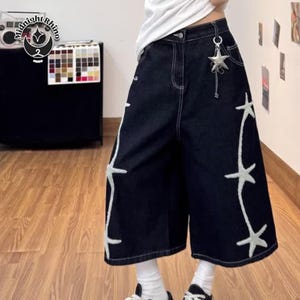 May include: Dark wash denim culottes with white star embroidery along the sides. The culottes have a high waist and a silver star charm attached to the belt loop. The pants are worn with white socks and black and white sneakers.