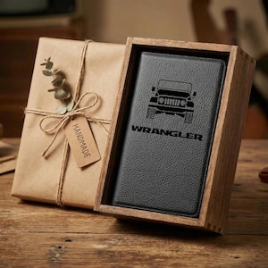 May include: A black leather wallet with the word "WRANGLER" and a Jeep graphic is displayed inside a wooden box. The box is open, revealing the wallet. The box is next to a wrapped gift with a "HANDMADE" tag.
