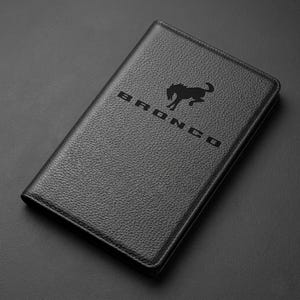 May include: Black leather-textured wallet with the word "BRONCO" and a horse logo embossed on the front. The wallet is rectangular and appears to be closed. The background is a dark gray surface.