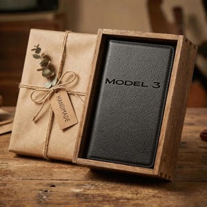 May include: A black leather Model 3 phone case in a wooden box. The case is embossed with the words "MODEL 3". The box is open, revealing the case. The box is sitting on a wooden surface. The box is next to a wrapped gift with a "HANDMADE" tag.