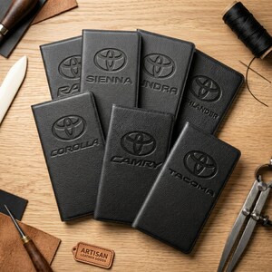 May include: Black leather Toyota vehicle key fob covers with embossed logos for various models, including Corolla, Camry, and Tacoma. The covers are arranged on a wooden surface, with artisan leather goods tools visible.