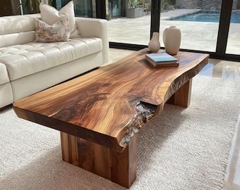 May include: A rectangular coffee table made from a single slab of natural wood with a live edge, featuring a prominent dark knot and visible grain patterns. The table is supported by two thick, solid wood block legs and has a smooth finish.